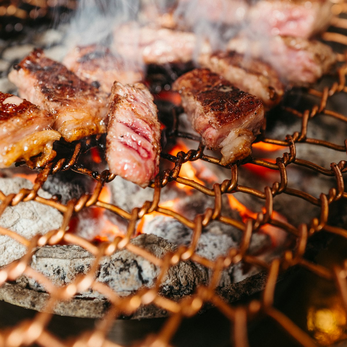 Smoke rising from thick cuts of beef searing on a grill over glowing charcoal